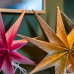 Close-up of decorative paper star ornaments hanging against a dark green wall, with one pink striped star on the left and a glittery gold star on the right.