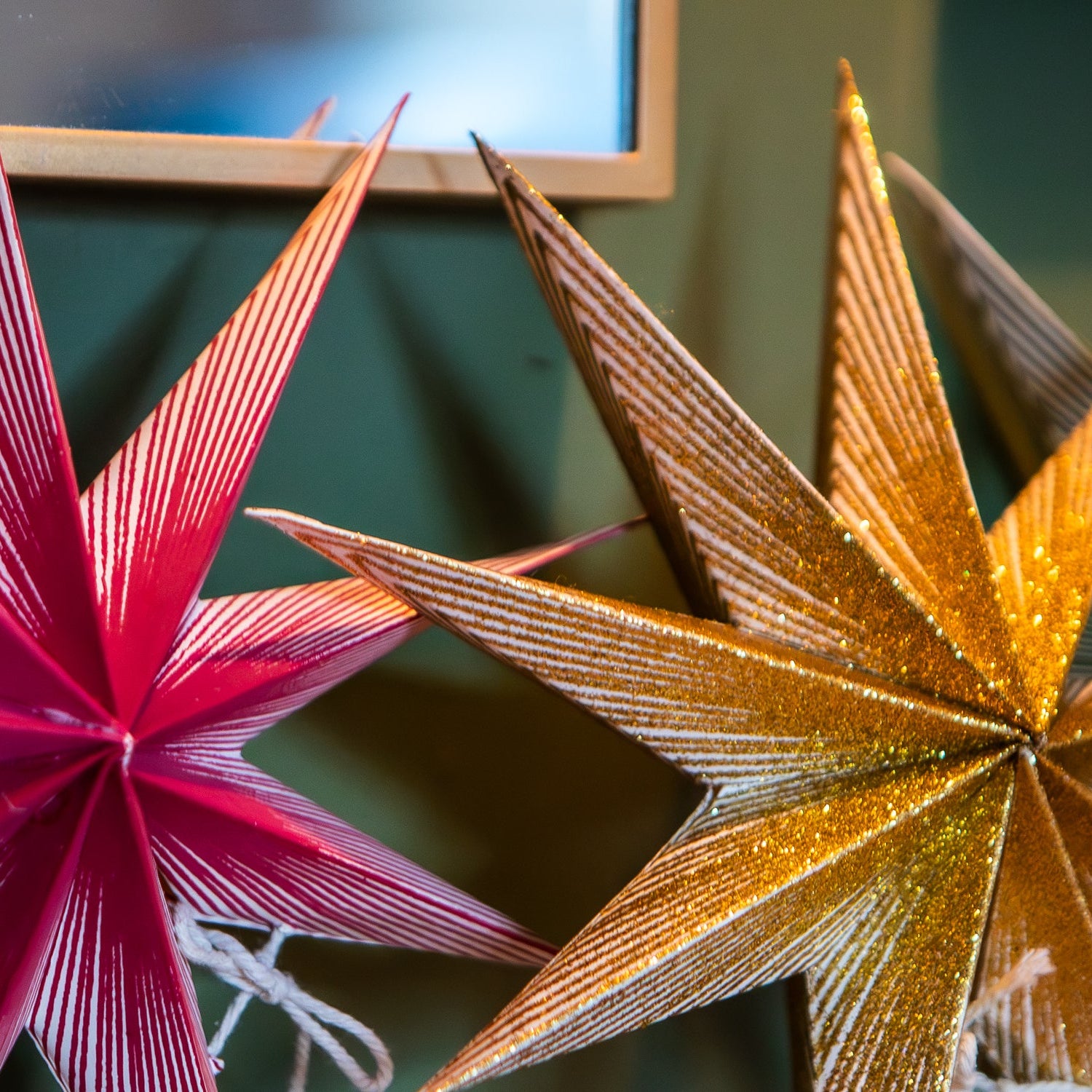 Close-up of decorative paper star ornaments hanging against a dark green wall, with one pink striped star on the left and a glittery gold star on the right.