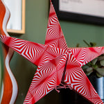 A red and white star-shaped decorative ornament with an optical illusion spiral pattern, shown hanging in a home setting next to a potted plant.