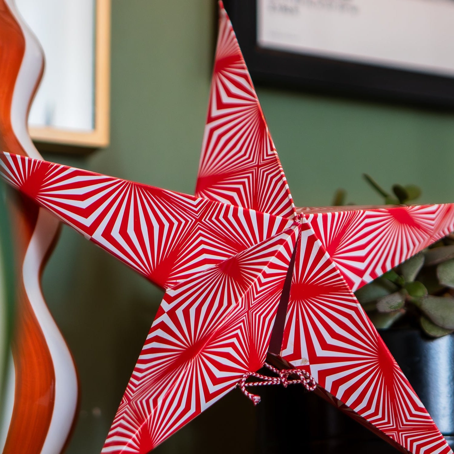 A red and white star-shaped decorative ornament with an optical illusion spiral pattern, shown hanging in a home setting next to a potted plant.