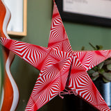 A red and white star-shaped decorative ornament with an optical illusion spiral pattern, shown hanging in a home setting next to a potted plant.