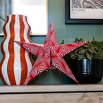 A red and white striped star-shaped decorative object displayed on a wooden mantel next to a wavy orange and white striped vase and a potted green plant against a green wall.