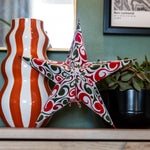 A decorative star-shaped ornament with a red, green, and white swirling pattern, displayed on a wooden shelf next to a wavy orange and white striped vase and a potted plant.