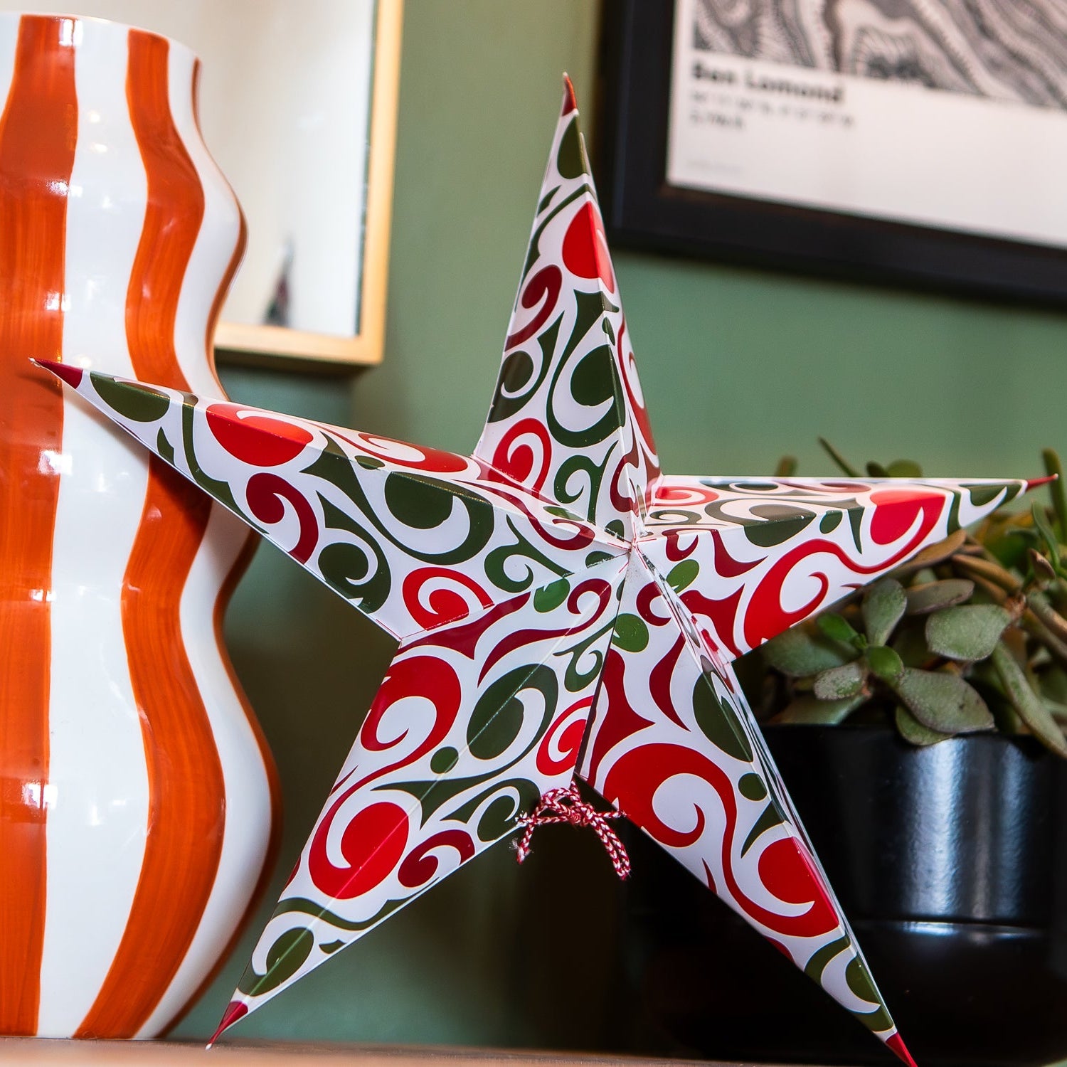 A decorative five-pointed star ornament with a red, green, and white swirling pattern, styled on a shelf next to a large orange and white striped vase and a potted succulent.