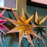 Three metallic star-shaped wall decorations hang on a dark green wall. The central star is gold, flanked by a pink star on the left and a bronze/gold star on the right.
