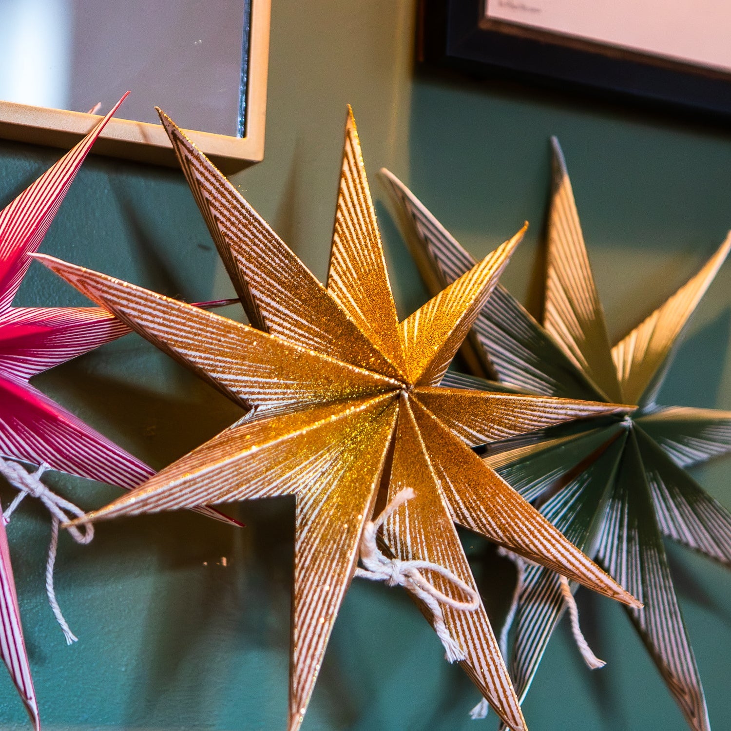 Three metallic star-shaped wall decorations hang on a dark green wall. The central star is gold, flanked by a pink star on the left and a bronze/gold star on the right.