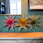 Three decorative paper star ornaments in pink, gold, and olive green hanging on a teal wall above a wooden mantel, with a mirror and framed print in the background.