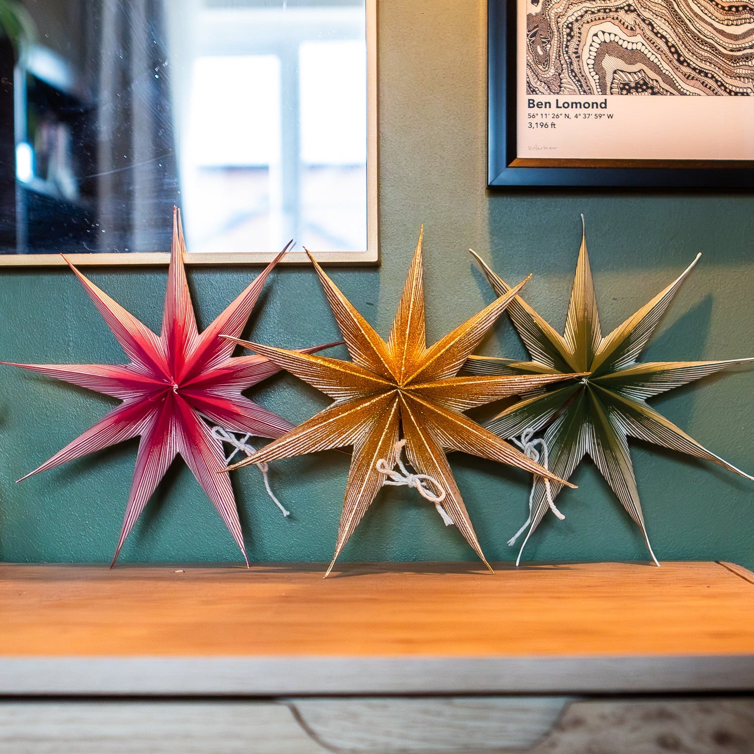 Three decorative paper star ornaments in pink, gold, and olive green hanging on a teal wall above a wooden mantel, with a mirror and framed print in the background.
