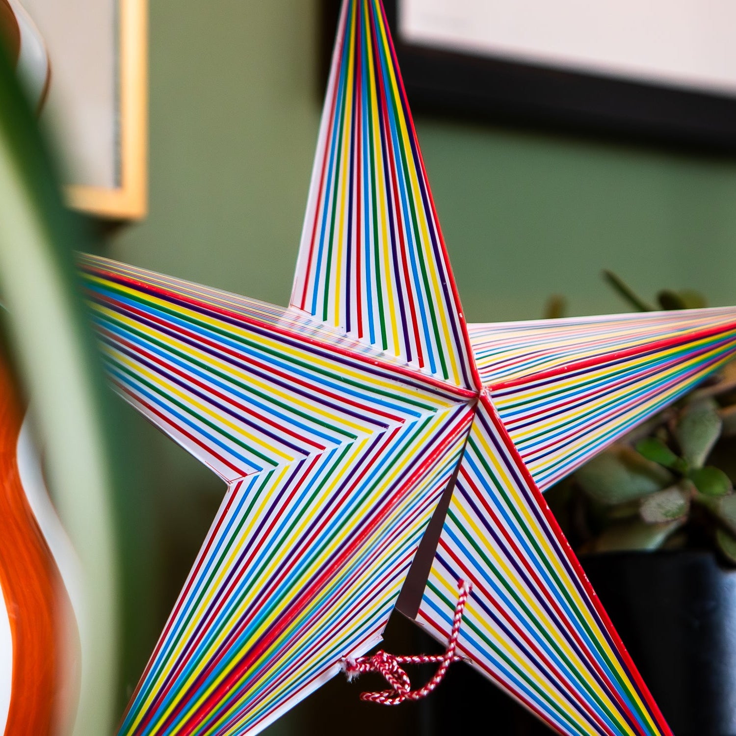 A decorative five-pointed star ornament made from narrow multicolored stripes, shown hanging indoors against a green wall with a plant and framed art in the background.