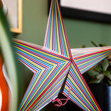 A decorative five-pointed star ornament made from narrow multicolored stripes, shown hanging indoors against a green wall with a plant and framed art in the background.