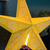 A close-up of a yellow five-point star decoration with white dotted circular patterns, photographed indoors against a dark teal background with blurred decor elements.