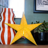 A yellow star-shaped decorative lantern with white mandala patterns stands on a wooden shelf next to a red and white striped ceramic vase and a potted green plant.