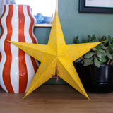 A bright yellow five-pointed star ornament with white circular patterns, standing on a wooden shelf next to a red and white striped vase and a potted green plant.