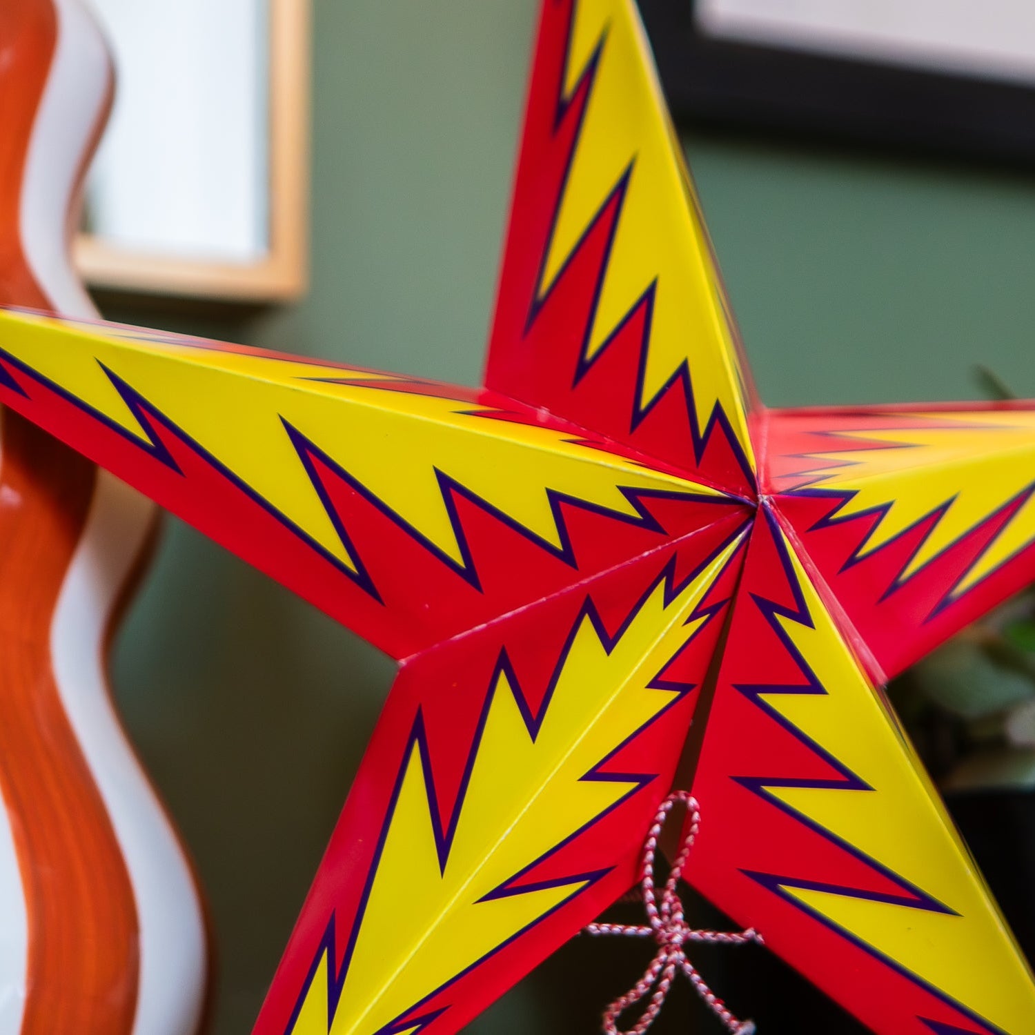 A close-up of a decorative five-point star ornament with alternating bright yellow and red sections outlined in dark blue, shown indoors against a blurred green wall and plant background.