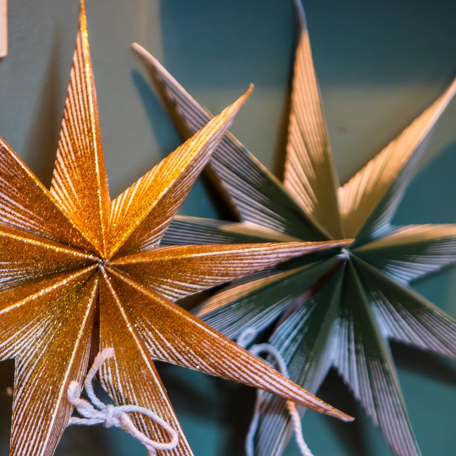 Two glittery paper star ornaments hanging on a dark teal wall, with one gold star in front and one green star behind it.
