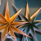 Two glittery paper star ornaments hanging on a dark teal wall, with one gold star in front and one green star behind it.