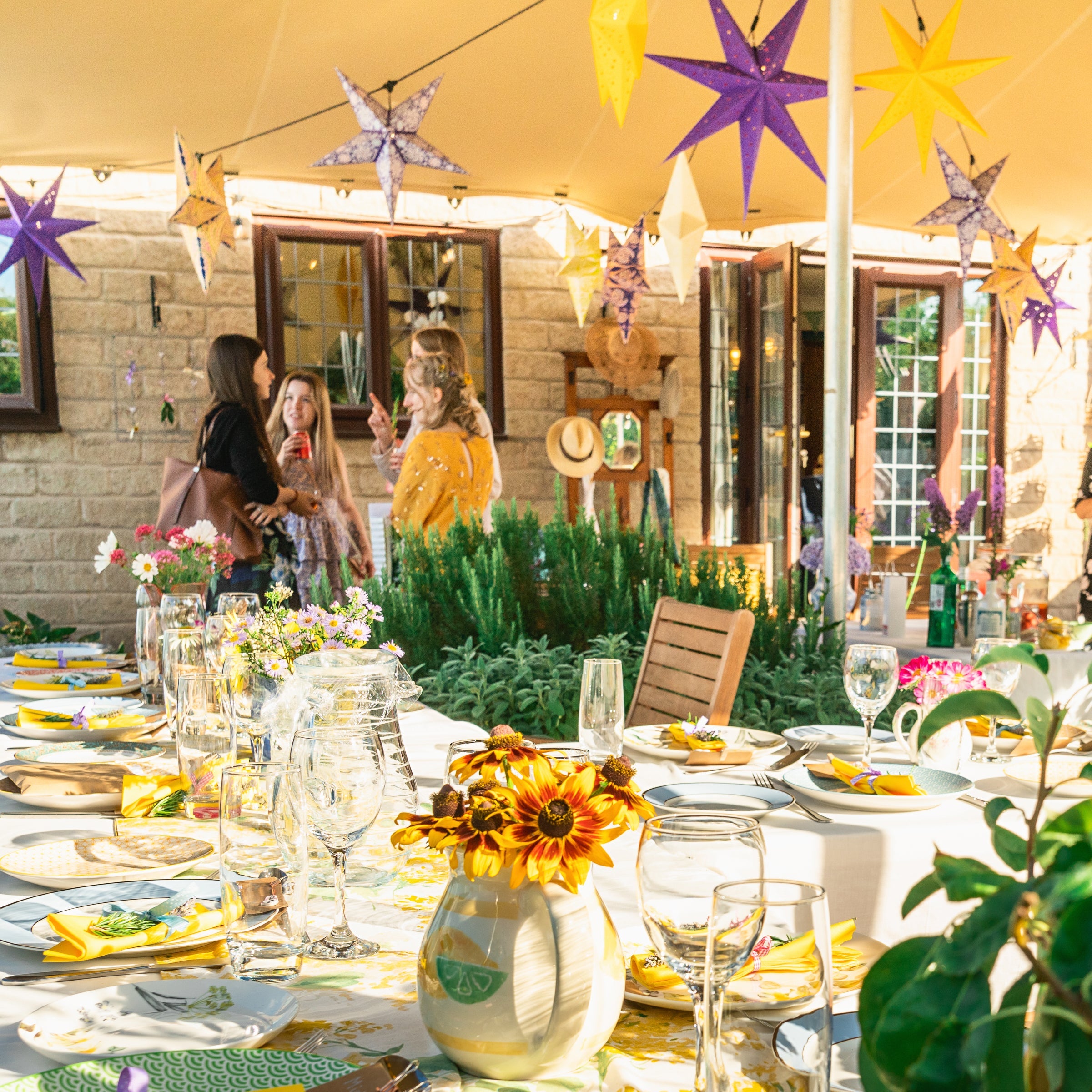 Outdoor table setting with yellow and white floral tablecloth, sunflowers, and guests in the background.